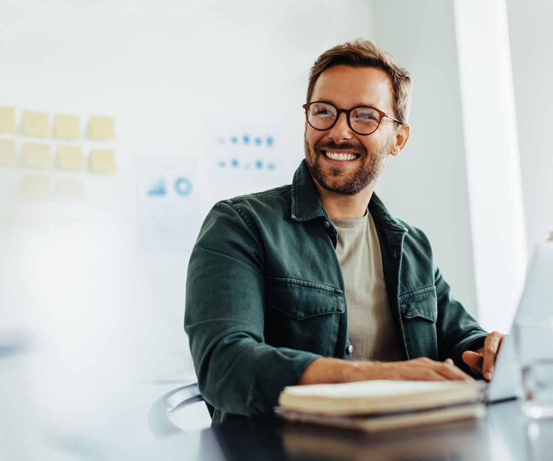 Lächelnder Mann mit Brille sitzt am Laptop im modernen Büro – Symbol für flexible und motivierende Lernformate bei MicroConsult.