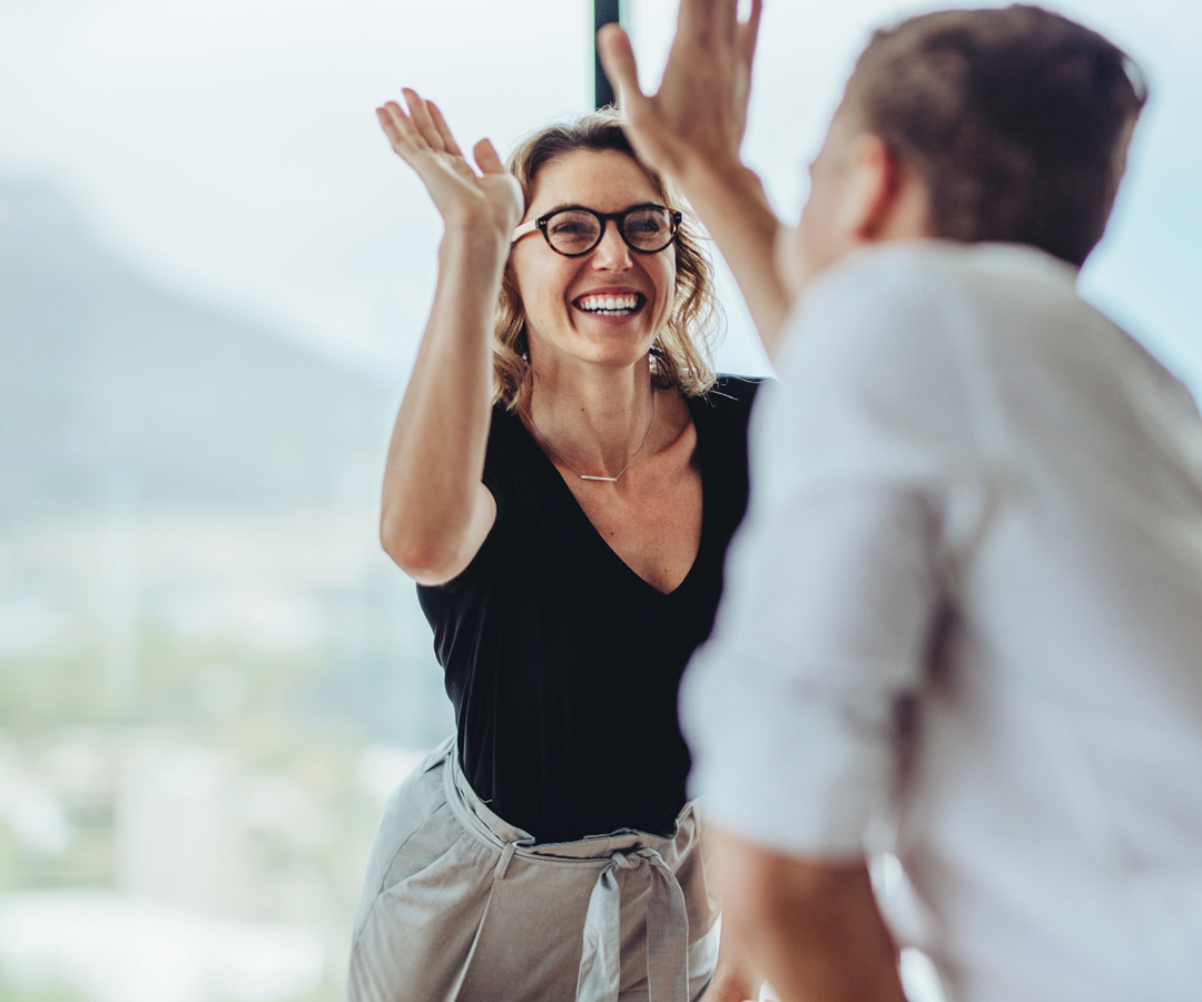 Zwei Kollegen geben sich ein High-Five im Büro, lachende Frau mit Brille im Vordergrund.