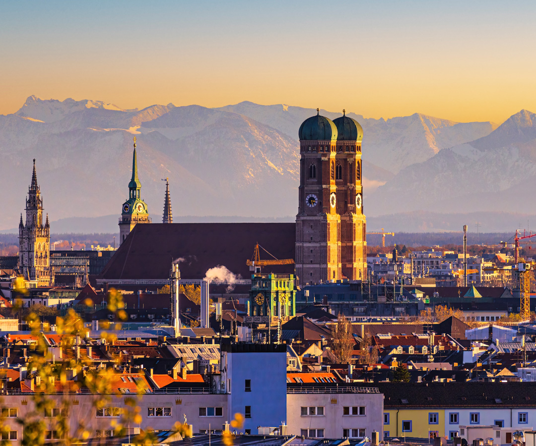 Panorama von München mit Frauenkirche, Marienkirche und Alpen im Hintergrund bei Sonnenuntergang.